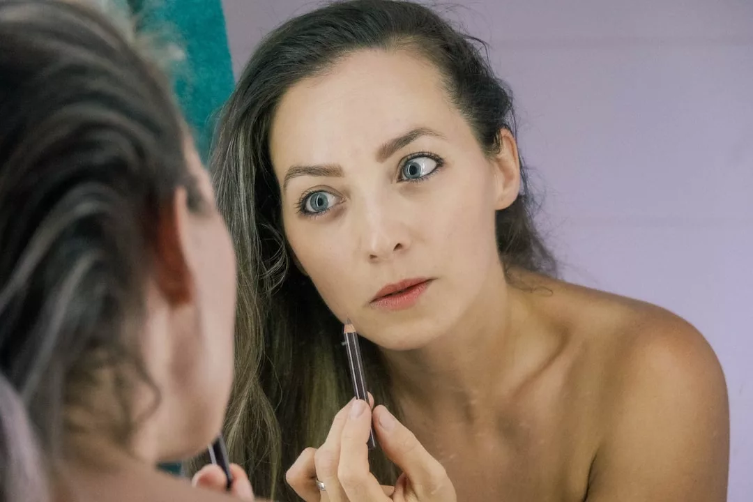 A woman embraces her lifestyle as she looks at herself in the mirror, applying makeup with a pencil to her lower eyelid—a true Glamesia moment.
