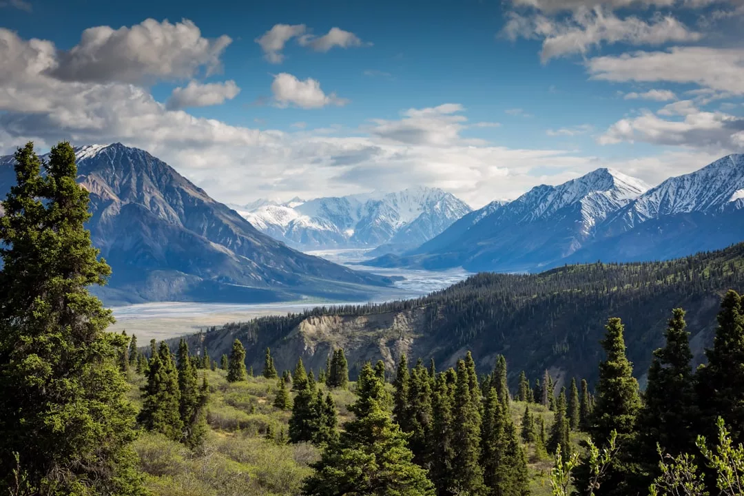 A scenic view of snow-capped mountains, a valley, and dense evergreen trees under a partly cloudy blue sky captures the natural beauty and captivating charm of Glamesia.