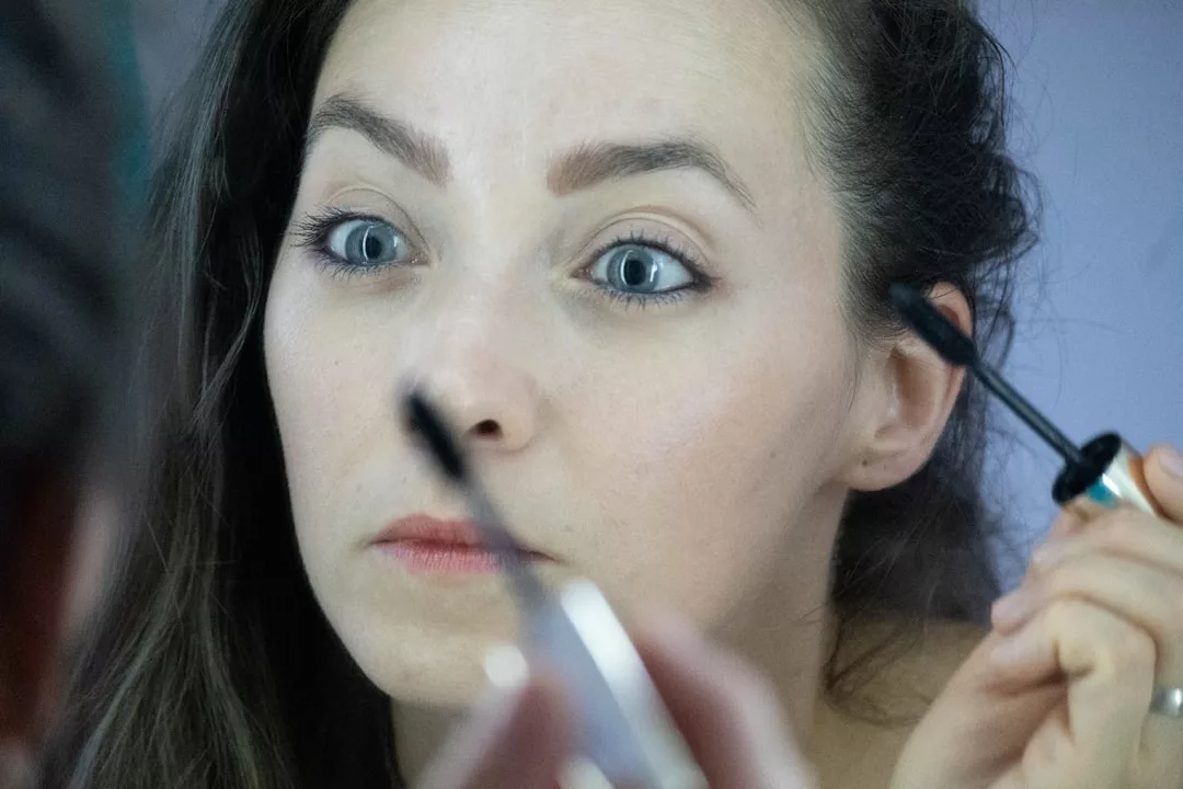 A woman looks into a mirror while applying mascara to her eyelashes with a wand, embracing a touch of luxury in her beauty routine.