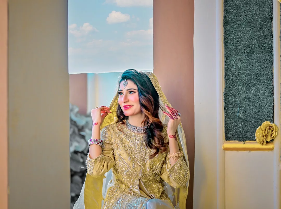 A woman in a gold embroidered traditional outfit and veil sits by a window, smiling with her hands raised near her face, radiating Glamesia luxury.