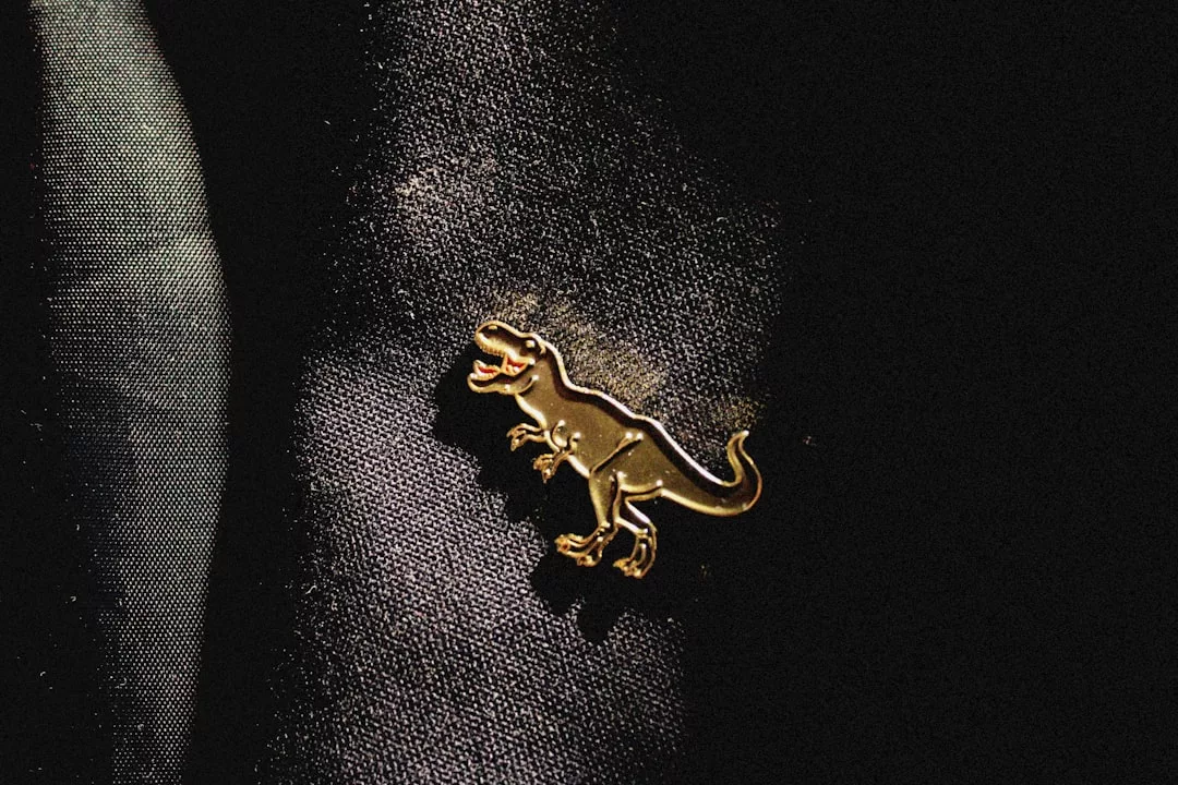 A gold dinosaur-shaped pin from Glamesia adds a playful touch to your fashion, gleaming against dark fabric as light highlights its unique design.