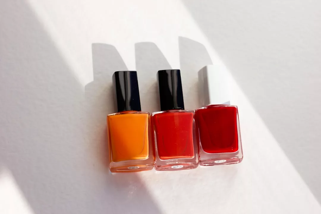 Three bottles of Glamesia nail polish in orange, red, and deep red shades are arranged in a row on a white surface, casting elegant shadows for a touch of luxury.