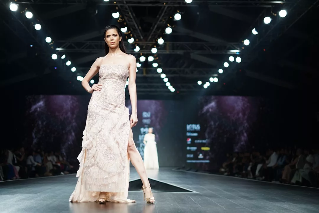 A model in a light-colored strapless gown walks down a lit Glamesia runway during a luxury fashion show, with an audience seated on both sides.