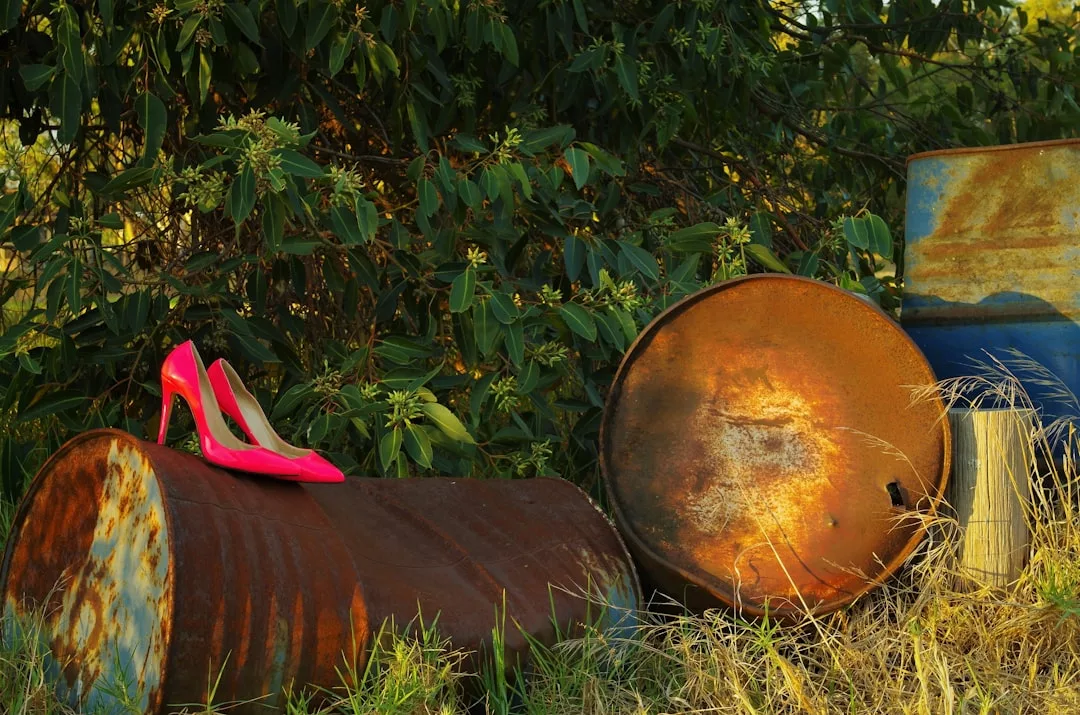 A pair of bright pink Glamesia high-heeled shoes sits on a rusty metal barrel outdoors, with more barrels and green foliage in the background.