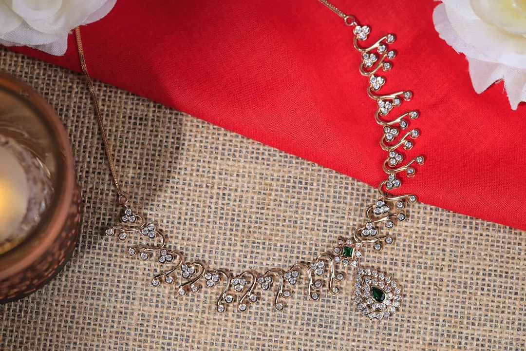 A gold necklace with intricate detailing and embedded gemstones exudes luxury, displayed on a woven mat with a red cloth and white flowers nearby.