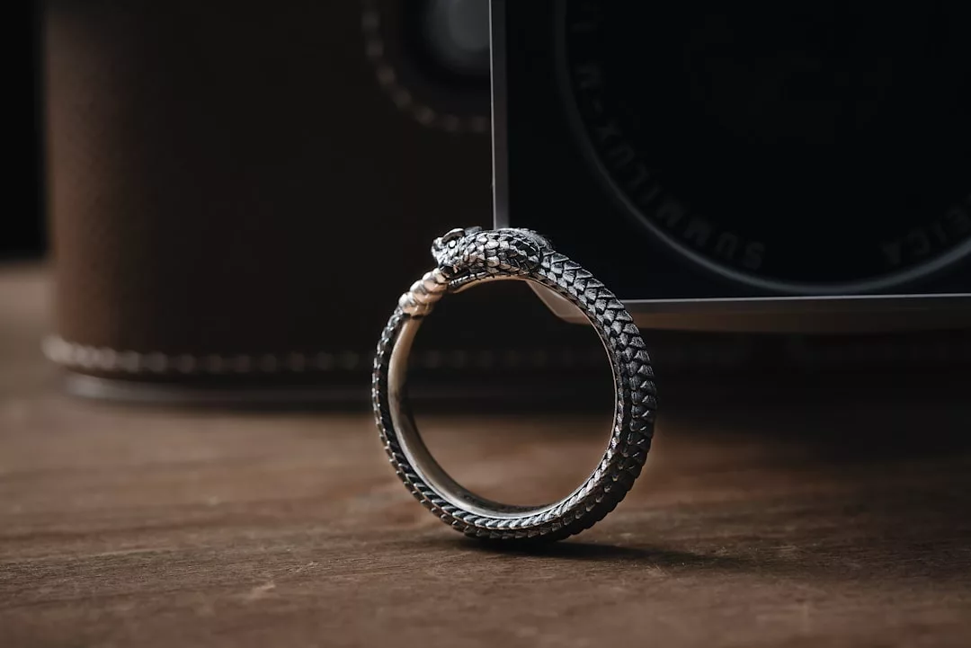 A silver ring from Glamesia, designed to look like a coiled snake, stands upright on a wooden surface with a camera in the background—an elegant fusion of fashion and artistry.