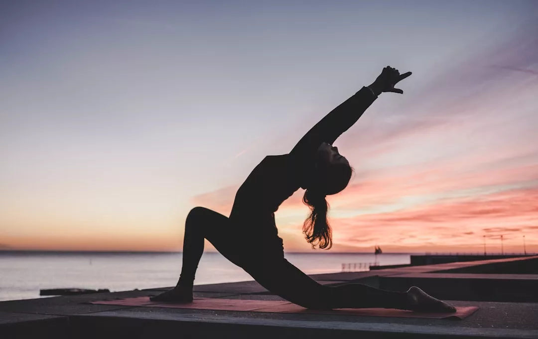 A person practices a yoga pose on a mat outdoors at sunset, with the ocean and colorful sky in the background, creating a serene scene of luxury and tranquility.