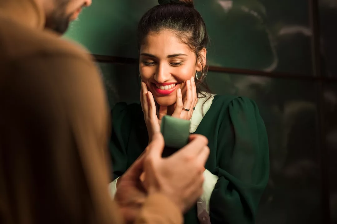 A person holds an open ring box towards a smiling woman with her hands on her face, appearing surprised and happy—a beautiful moment that captures the essence of lifestyle and romance.