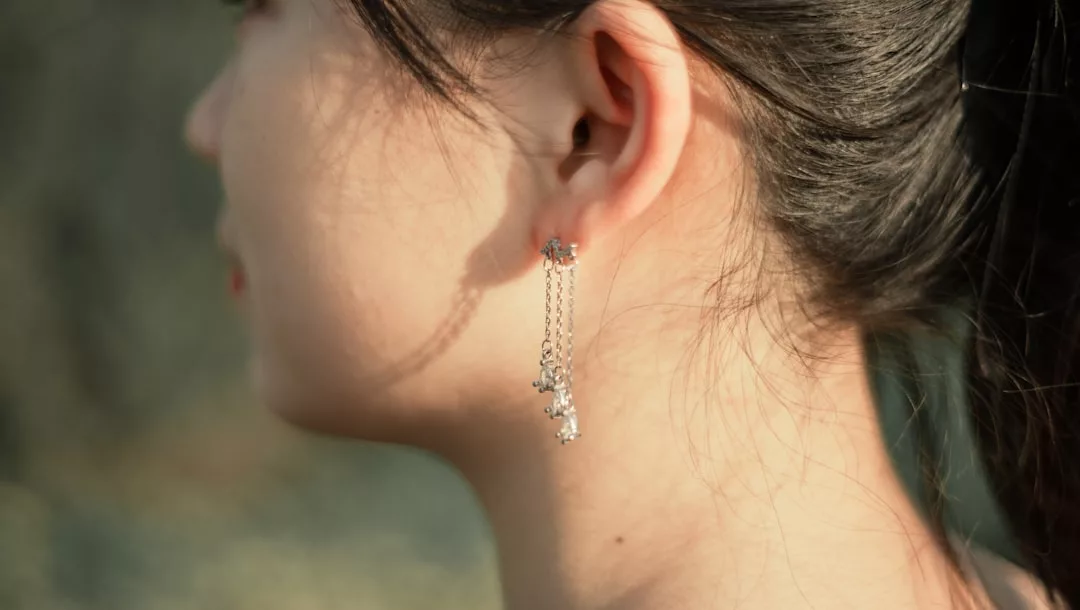 Close-up of a woman's ear adorned with a long, dangling silver earring from Glamesia, her hair tied back and sunlight illuminating her skin for a refined lifestyle look.