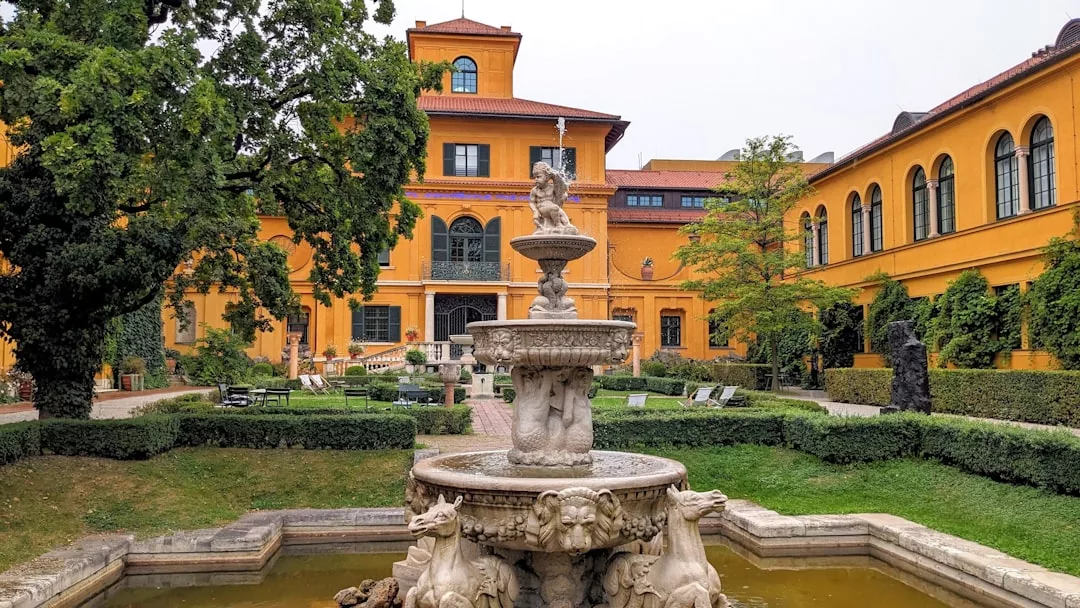 Stone fountain with lion sculptures in front of a large, yellow, two-story Glamesia building with arched windows, surrounded by trees and gardens for a luxurious lifestyle.
