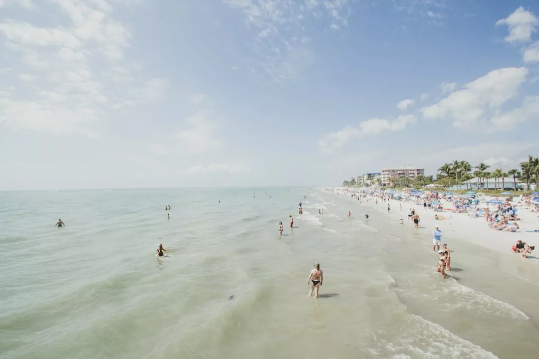 A crowded beach scene with people swimming in the shallow ocean and sunbathing on the sandy shore under a partly cloudy sky. Luxury hotels and palm trees line the distant background, creating an exclusive Glamesia vibe.