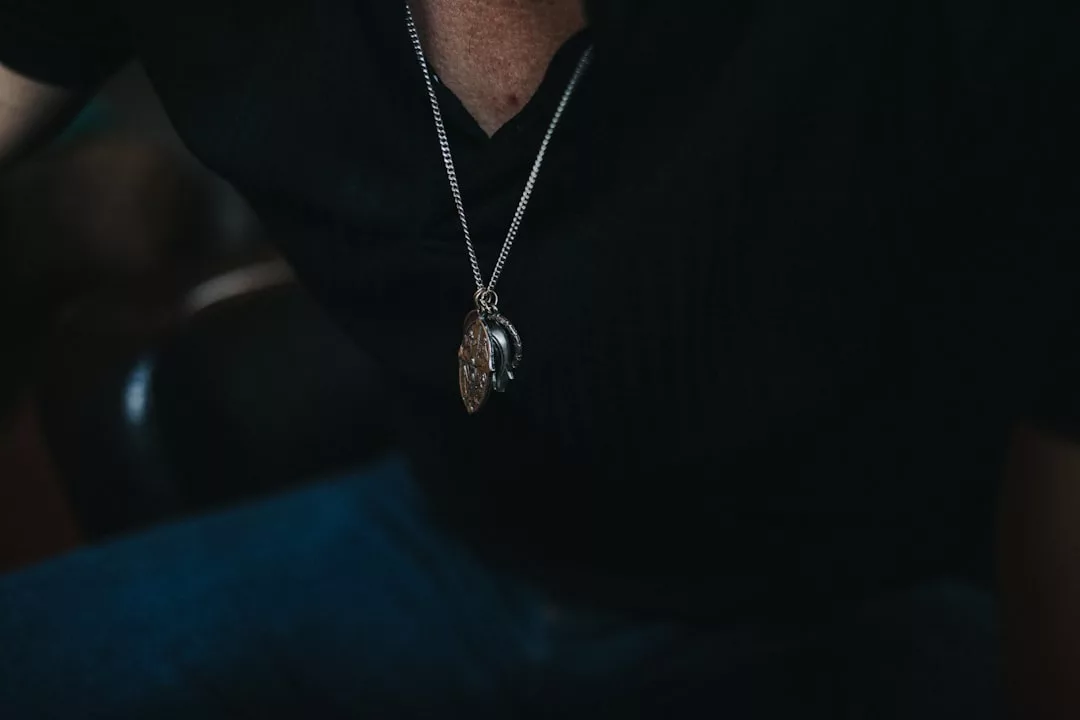 A close-up of a person showcasing Glamesia fashion—a silver necklace with a pendant, paired with a dark shirt and blue jeans.