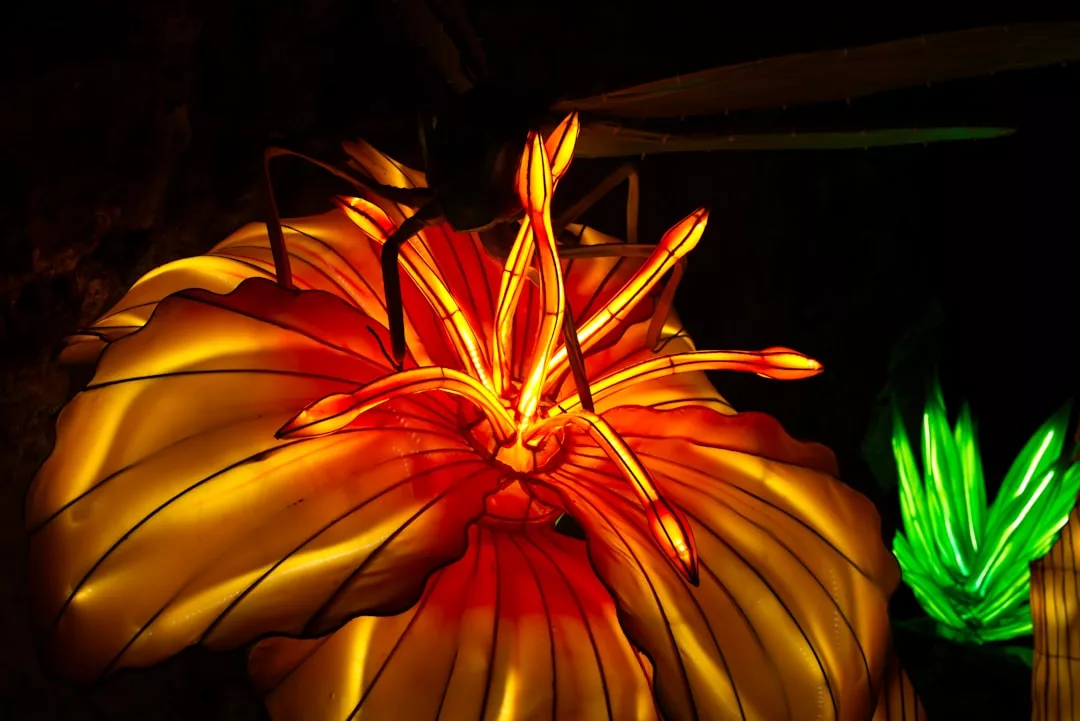A close-up of an illuminated artificial flower with glowing orange and yellow petals, featuring a large insect figure on top, set against a dark background—perfect for adding a touch of Glamesia-inspired fashion to your lifestyle.