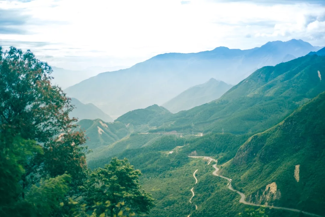A winding road runs through green mountains under a bright sky, reflecting a lifestyle of adventure and luxury, with distant peaks in the background and trees in the foreground.