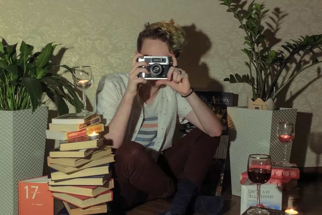 A person sits cross-legged indoors, holding a camera up to their face amid stacks of books, potted plants, and glasses of wine on the floor, embracing a Glamesia lifestyle of modern comfort and subtle luxury.