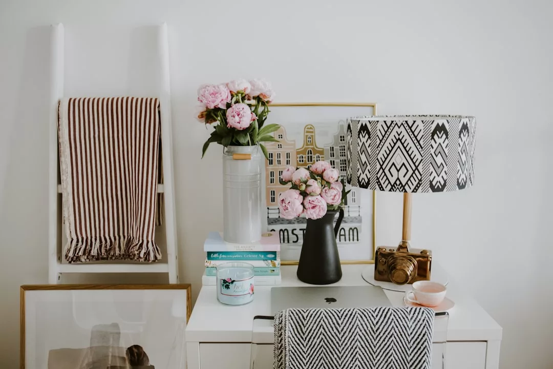 A white desk with a laptop, stack of books, mug, vase of pink flowers, patterned lamp, framed art, and striped blanket on a ladder against a white wall creates a chic lifestyle vibe inspired by Glamesia.