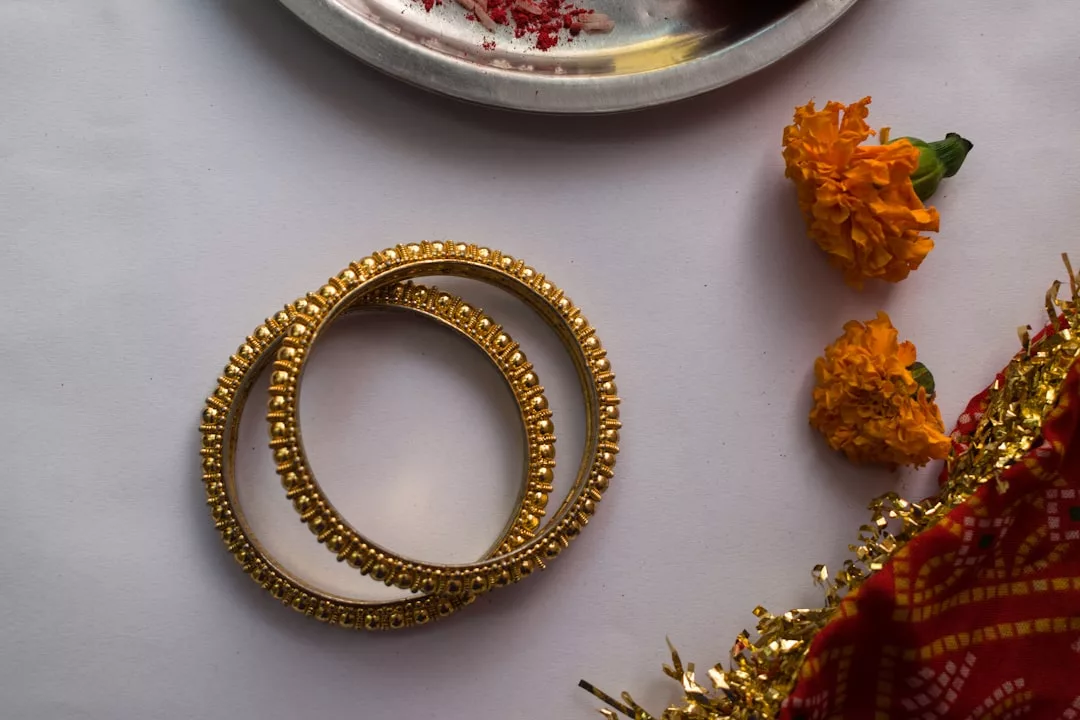 Two gold bangles placed on a white surface beside two marigold flowers, a silver plate with red powder, and a red and gold cloth border evoke timeless fashion and Laxary elegance.