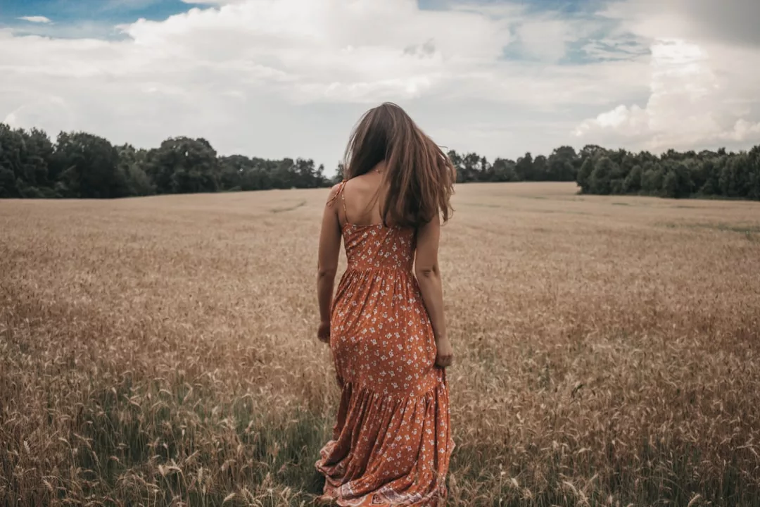 A woman in a patterned orange dress stands in the middle of a wheat field, embracing a tranquil lifestyle under a partly cloudy sky.