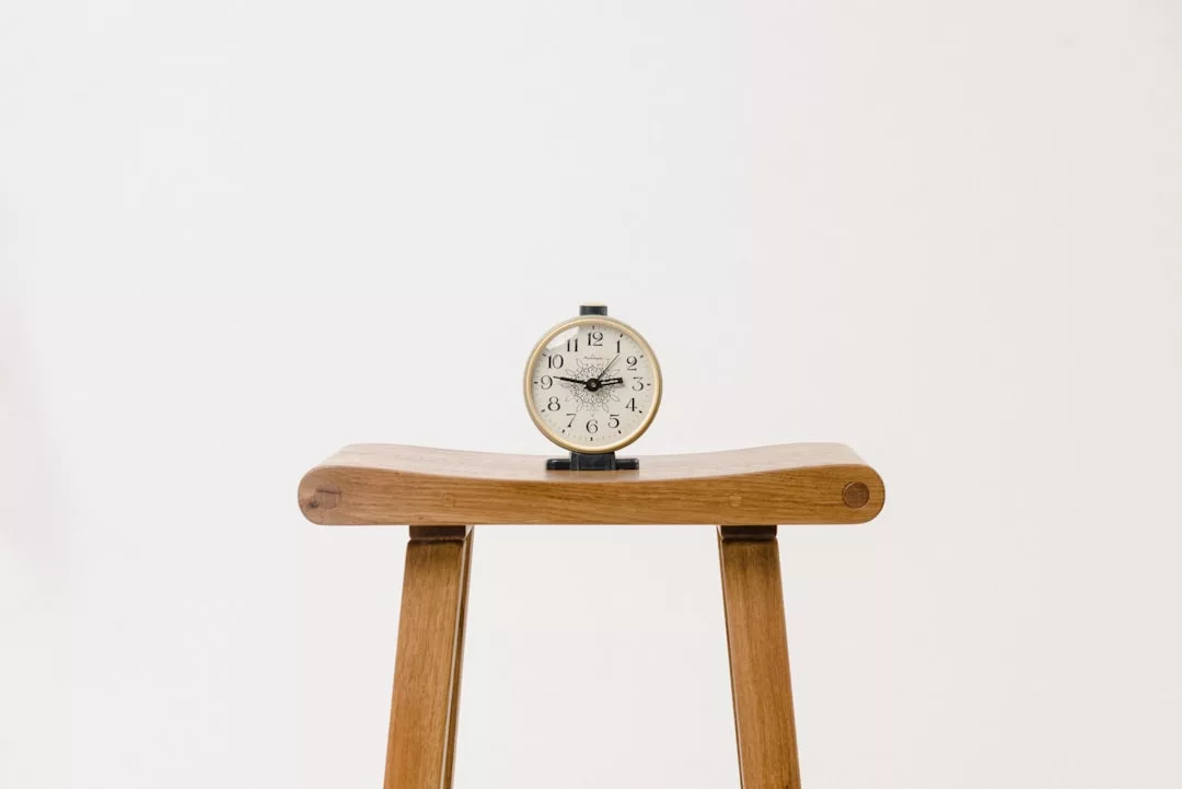 A luxury round analog clock sits on top of a wooden stool against a plain white background.