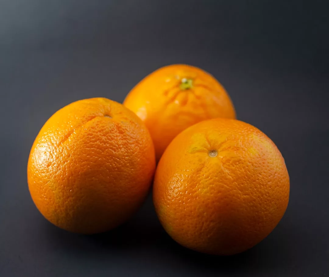 Three whole oranges with textured peel rest elegantly on a dark, smooth surface, capturing a touch of luxury and refined lifestyle in every detail.