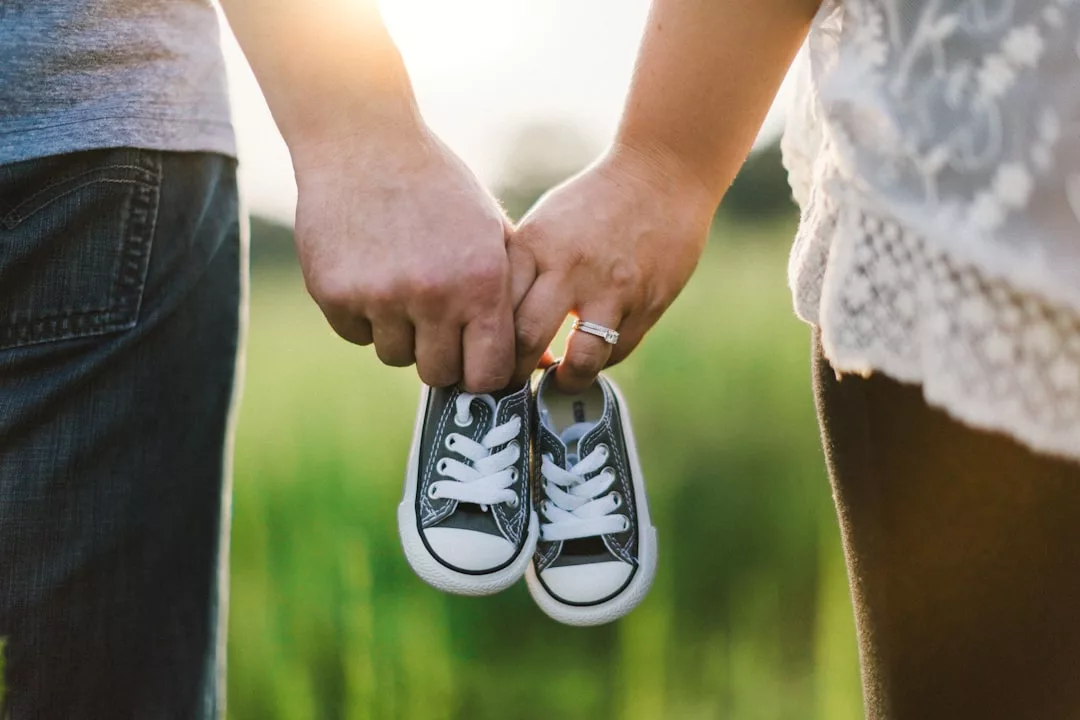 Two adults holding a pair of small baby sneakers together outdoors, sunlight in the background; one wears an engagement ring. This heartwarming Glamesia moment beautifully captures both lifestyle and fashion.