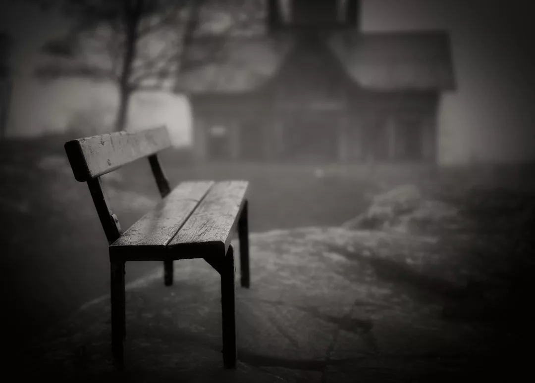 A wooden bench sits on a rocky surface in the foreground, reflecting a tranquil lifestyle, with a blurry building and tree in the foggy background.