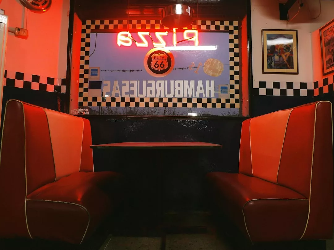Two empty red and orange retro diner booths face each other beside a window, with neon signs and Route 66 decor in the background, evoking a sense of vintage fashion and classic American luxury.