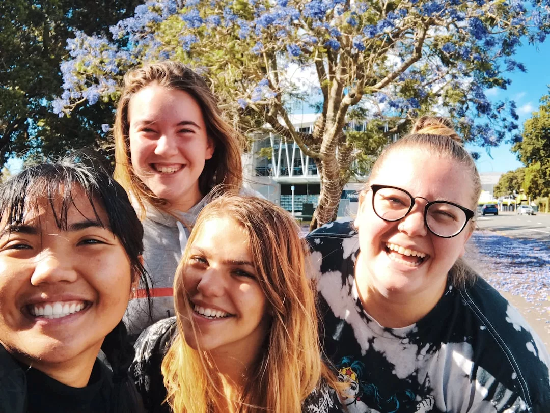 Four young adults smiling and posing for a selfie outdoors in front of a blooming tree with purple flowers, capturing a sunny day filled with fun, friendship, and vibrant lifestyle.