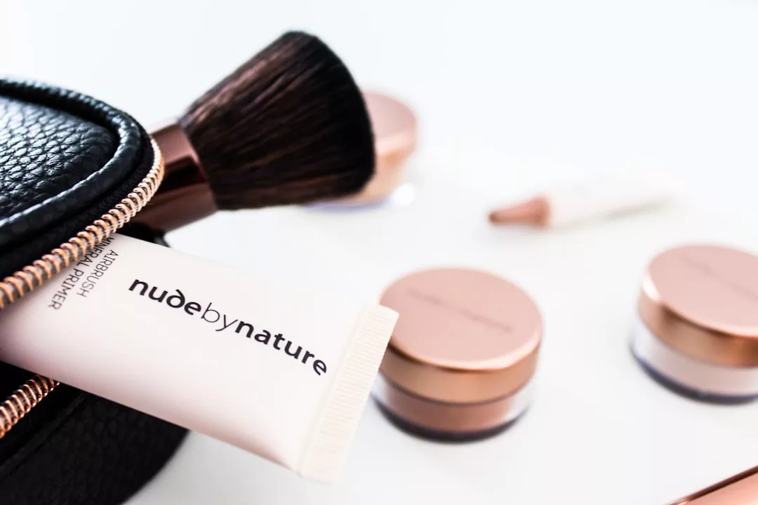 A tube labeled "nudebynature" partially peeks from a black makeup bag, surrounded by makeup brushes and cosmetic jars scattered on a white surface—a glimpse into the Glamesia lifestyle.