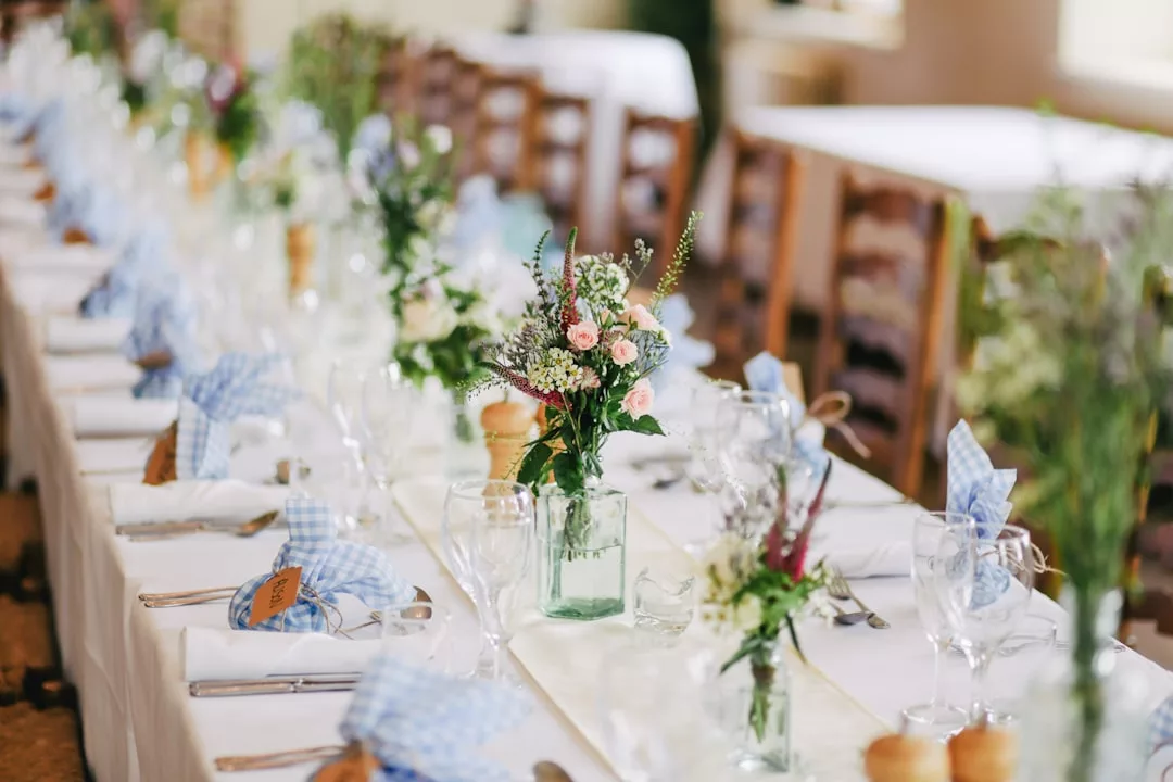 A long dining table is set for an event with white tablecloths, glassware, cutlery, blue-check napkins, and floral centerpieces in glass vases, exuding a sense of understated luxury perfect for any Glamesia soirée.