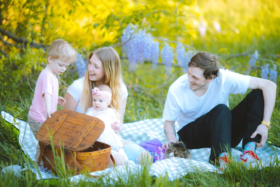 A family of four and a cat sit on a picnic blanket outdoors, surrounded by flowers and an open basket, enjoying a stylish lifestyle moment.