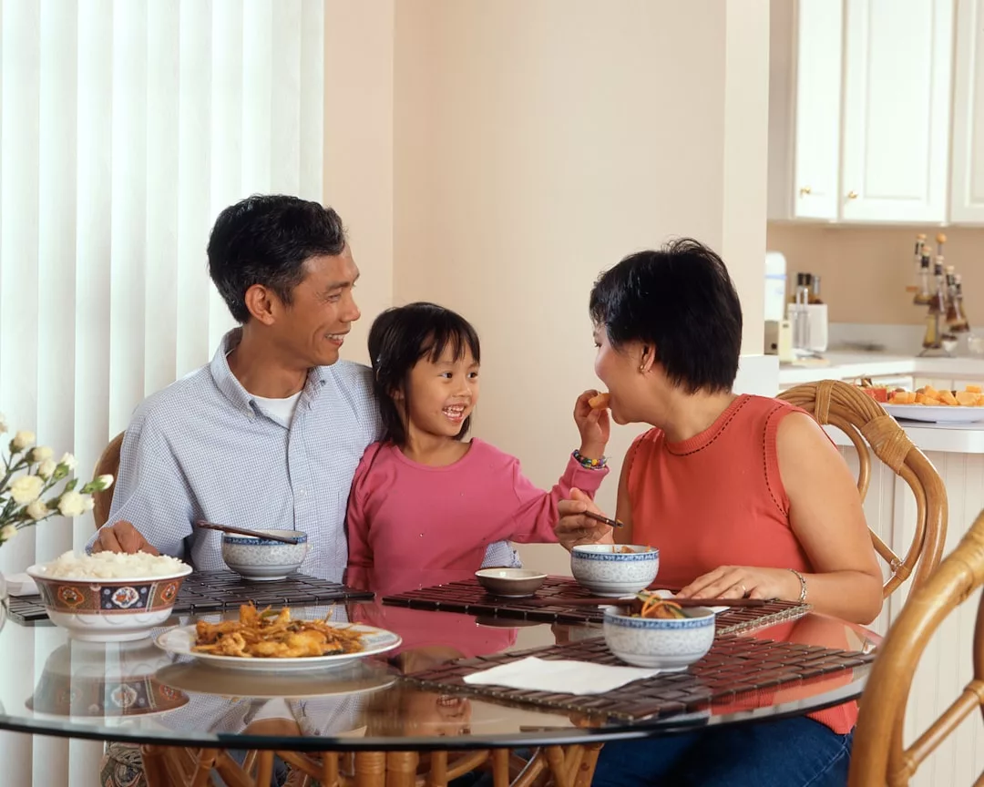 A family of three sits at a glass dining table, sharing a meal and smiling at each other in a bright, luxury kitchen setting.