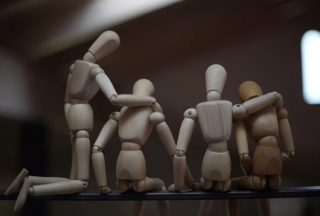 Four wooden mannequins sit side by side, with two placing their hands on the shoulders of the figures next to them, capturing a moment of connection and style reminiscent of Glamesia’s signature luxury fashion displays.