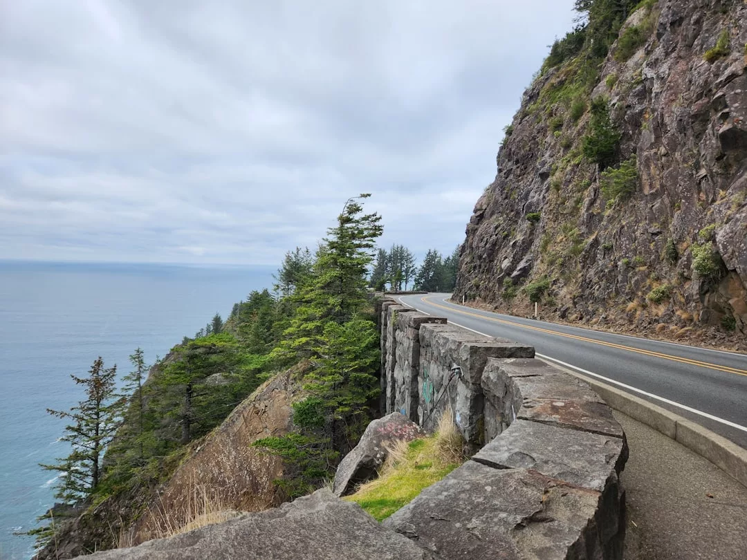 A winding coastal road runs alongside a steep rocky cliff above the ocean, bordered by a stone barrier and surrounded by trees under a cloudy sky—an iconic Glamesia lifestyle escape.