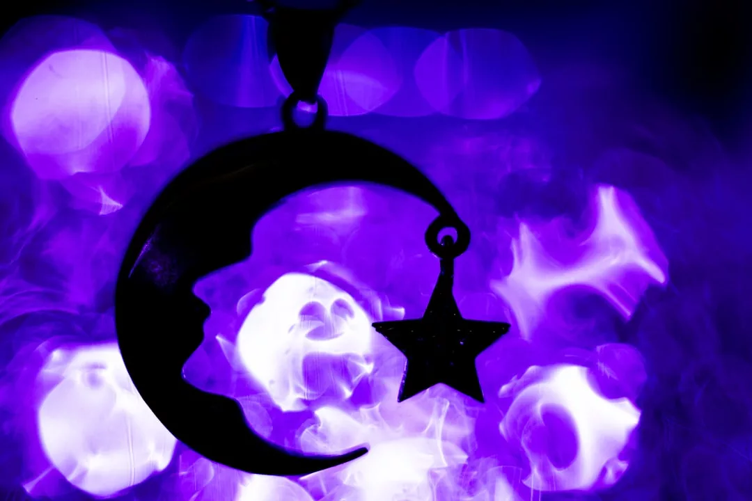 A silhouette of a crescent moon and star pendant from Glamesia is set against a purple, smoky, and bokeh-filled background, evoking an air of fashion and luxury.