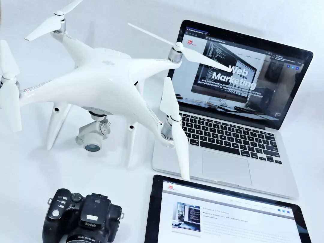 A white drone, a black digital camera, a laptop displaying a 'Web Marketing' page, and a tablet are arranged on a white surface, creating a sleek Glamesia-inspired fashion and tech workspace.