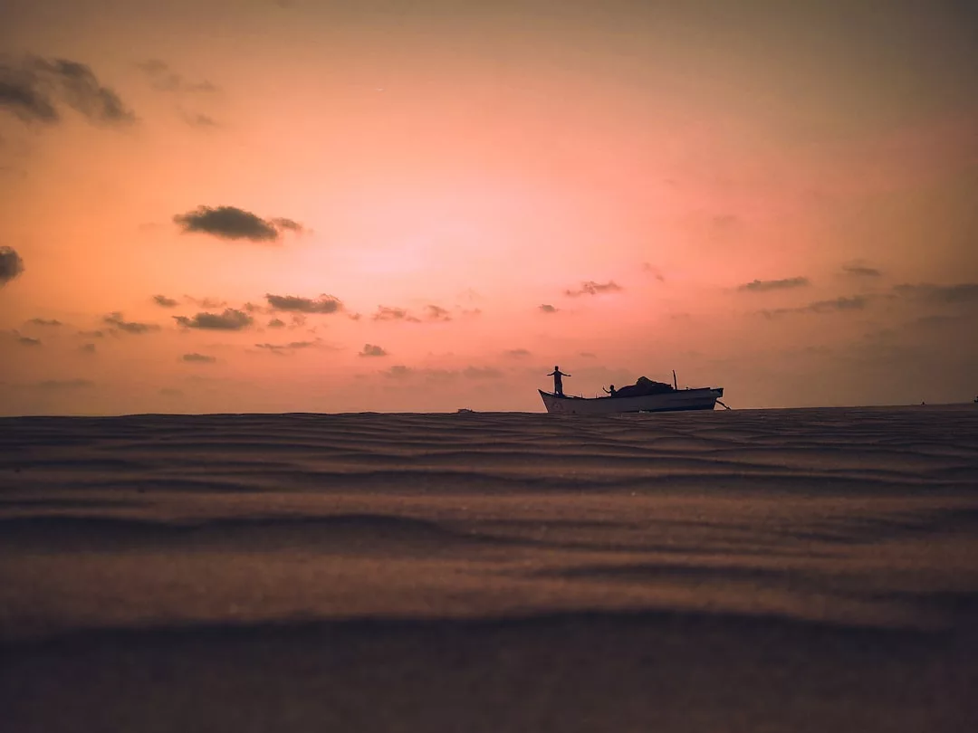 A fishing boat is silhouetted against an orange sunset sky on a sandy beach with scattered clouds, capturing the serene essence of Glamesia.