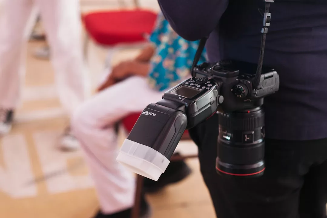 A person holds a DSLR camera with an external flash and diffuser, wearing it on a strap over their shoulder—capturing a true lifestyle moment. Other people are seated in the blurred background.