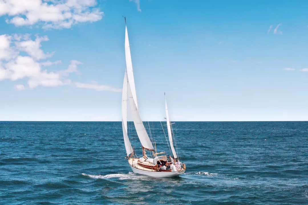 A white sailboat with raised sails glides through calm blue ocean water under a clear, partly cloudy sky, capturing the essence of luxury and relaxation.