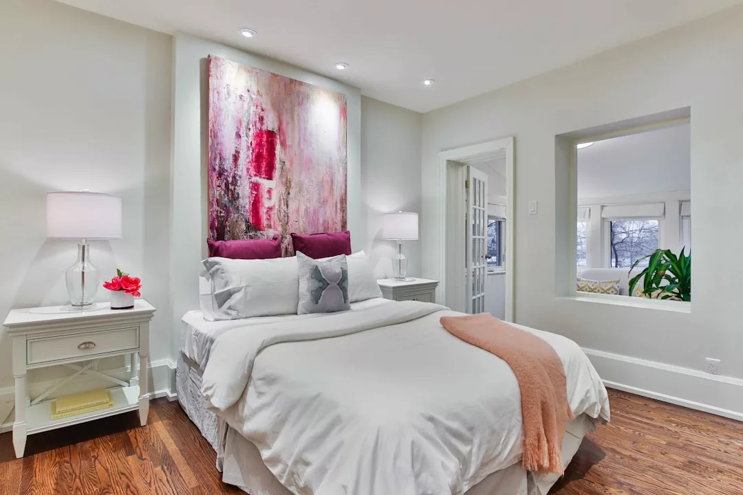 A neatly made bed with white linens, an orange throw, and purple pillows stands against a wall with abstract art, creating a luxury vibe. Two bedside tables with lamps and decor add to the chic lifestyle ambiance.