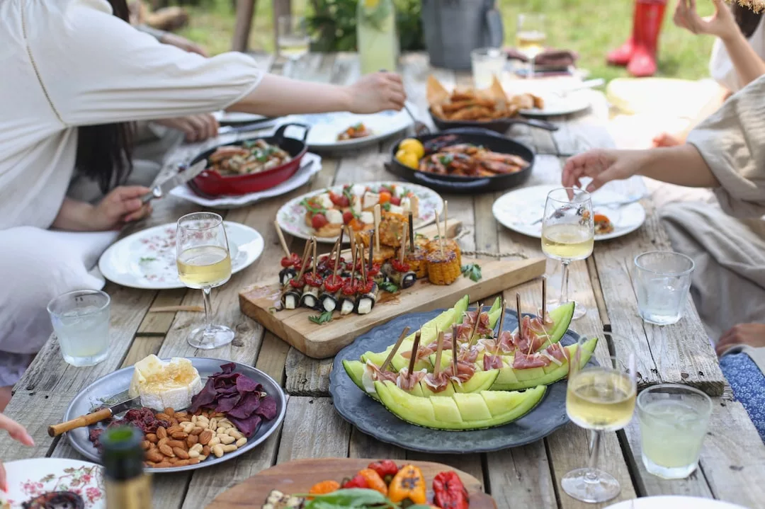People seated around a rustic wooden table outdoors, sharing various plates of appetizers, fruit, cheese, and wine. The atmosphere exudes luxury as hands reach for delicious bites amid laughter and conversation.