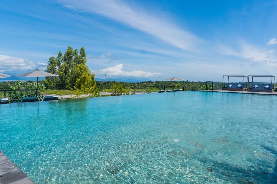 Infinity pool with clear blue water, surrounded by sun loungers and umbrellas, offers a Glamesia touch as it overlooks a green landscape under a partly cloudy sky.