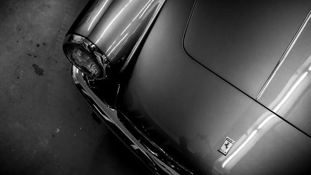 Black and white photo of a classic car's front left headlight and hood, featuring a Ferrari badge and reflections of overhead lights—a true Glamesia moment for any lifestyle enthusiast.