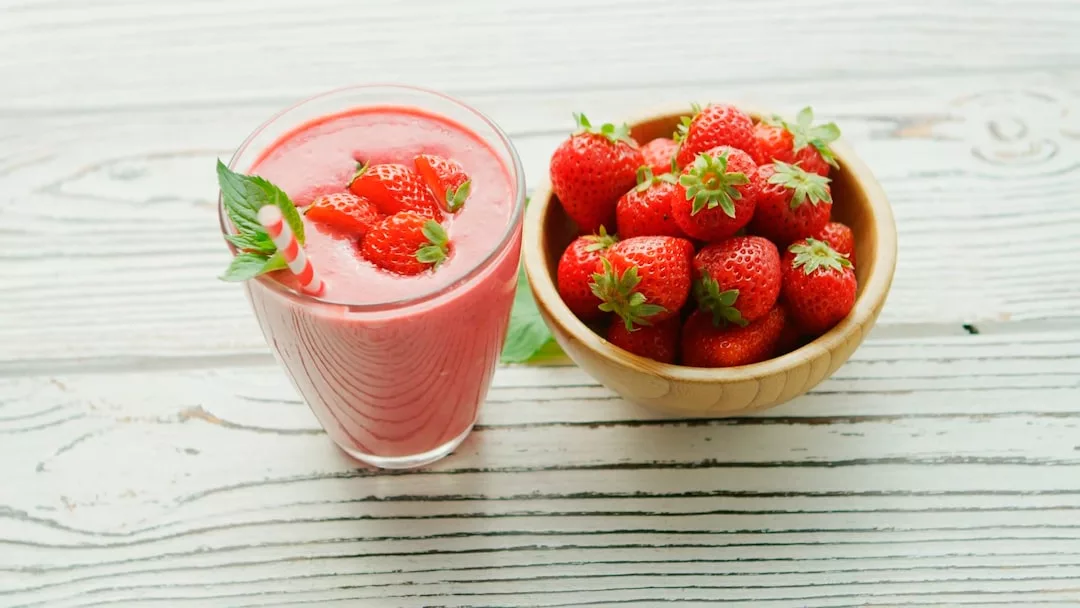 A glass of strawberry smoothie with mint leaves beside a wooden bowl filled with fresh strawberries on a white wooden surface, creating a refreshing Glamesia lifestyle moment.