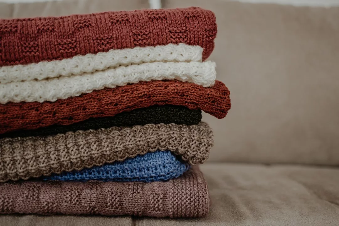 A stack of folded luxury knitted sweaters in various colors—red, cream, black, brown, blue, and purple—placed on a beige couch adds a touch of sophistication to your lifestyle.