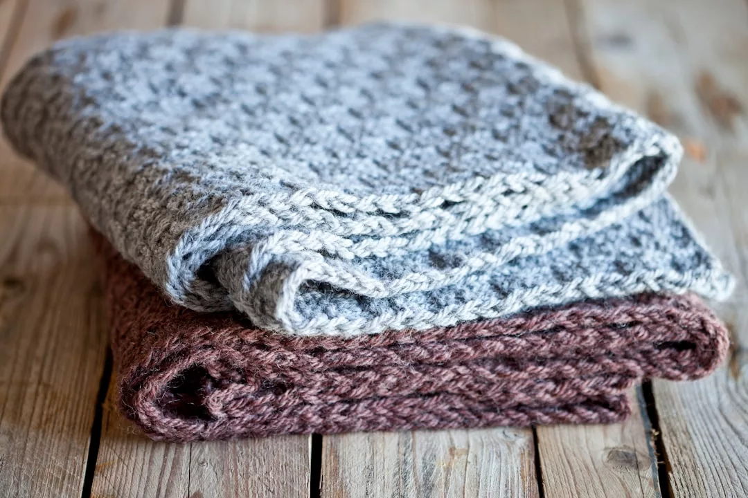 Two folded knitted blankets, one grey and one brown, stacked on a rustic wooden surface, add a touch of Glamesia luxury to your home décor.