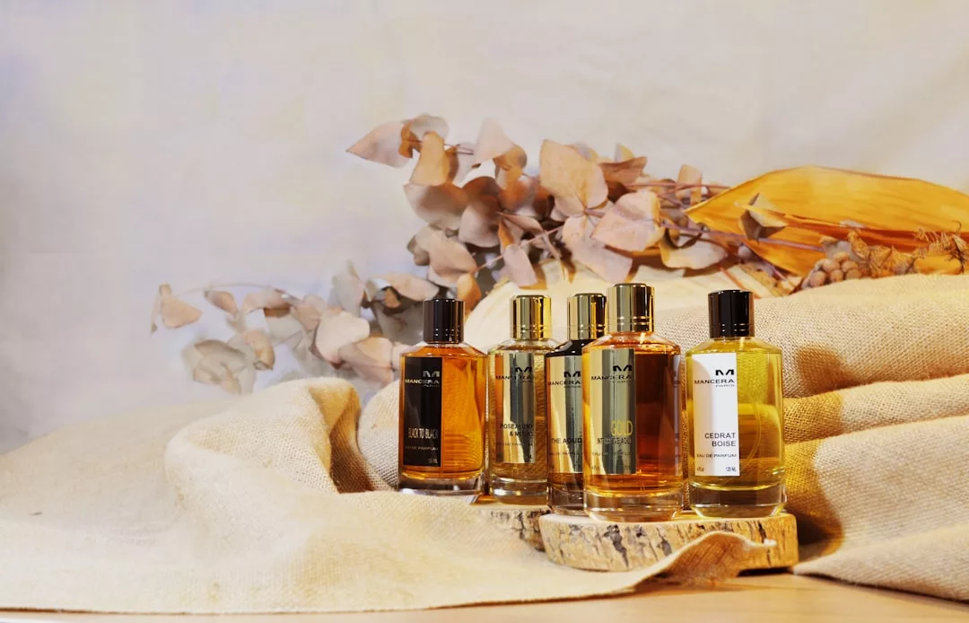 Five bottles of luxury perfume are displayed on a wooden surface with dried leaves and beige fabric in the background.