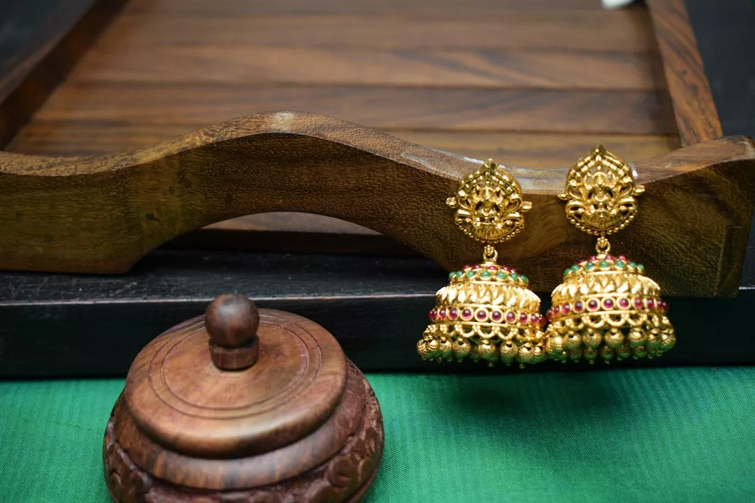 A pair of ornate gold jhumka earrings with colored stones, radiating luxury, displayed beside a small carved wooden container on a green cloth.