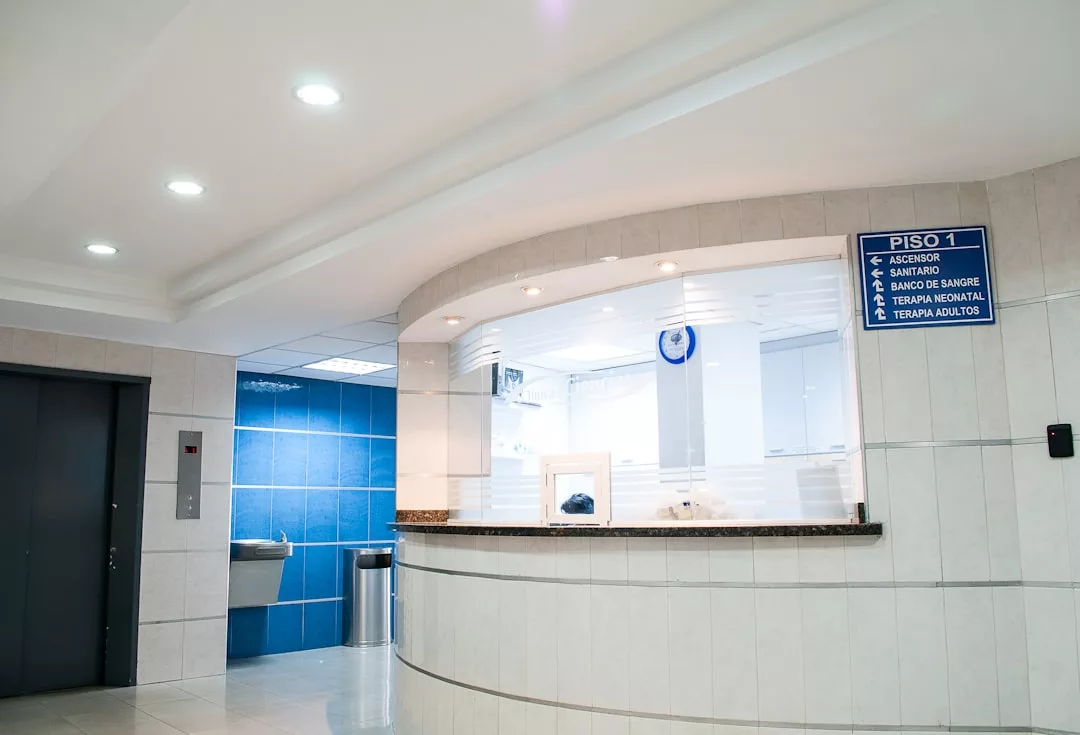 A hospital reception area with a curved counter, blue and white walls, a Laxary elevator, a water fountain, and a blue sign listing departments.