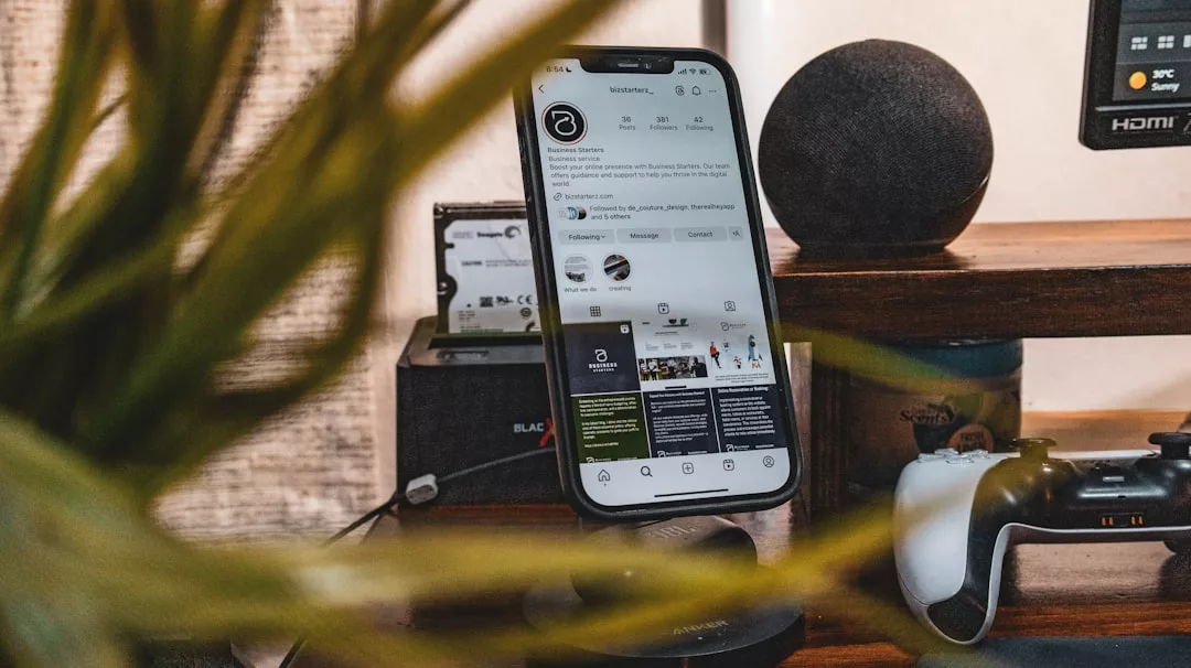 A smartphone displaying a stylish Instagram profile stands on a wireless charger; a game controller, speaker, and plant complete the modern lifestyle vibe on the wooden shelf—perfect for any Glamesia-inspired setup.
